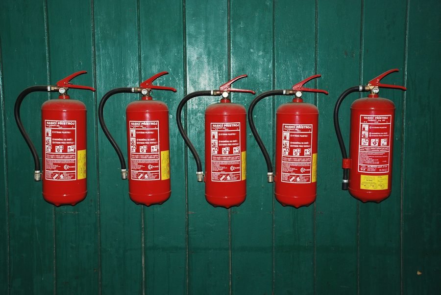 Five red extinguishers hanging in a row on a dark green painted wooden wall.